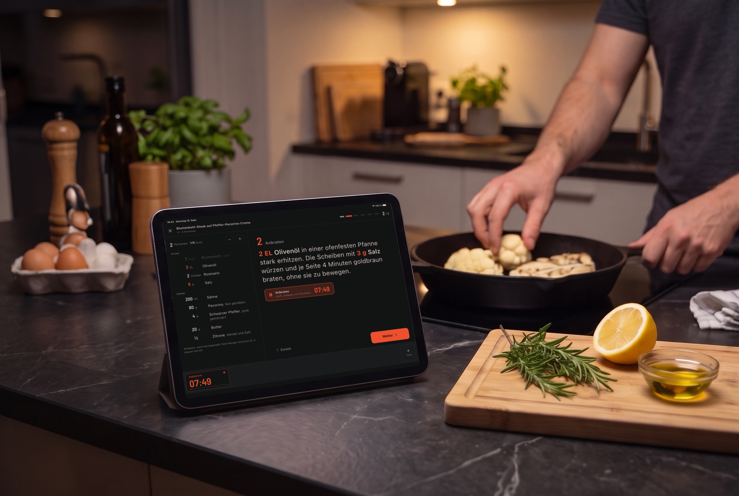 Evening in the kitchen: a tablet on the counter shows the cook mode of Cook & Repeat, and behind it someone turns cauliflower in a cast-iron pan.